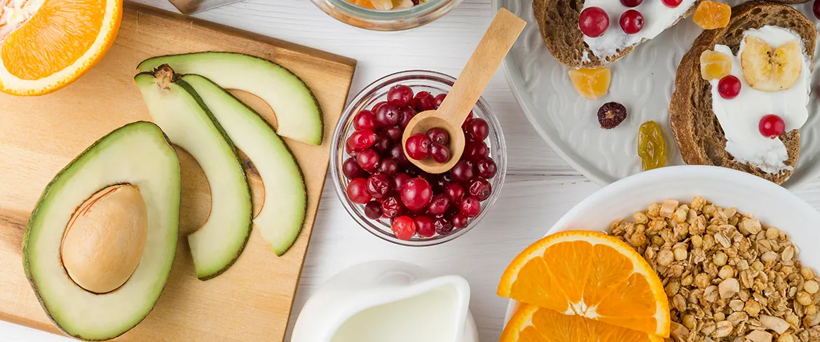 Różnorodne zdrowe produkty śniadaniowe, w tym pokrojone awokado na drewnianej desce, miska z żurawinami, jogurt z owocami na toście, granola w misce i pokrojone pomarańcze